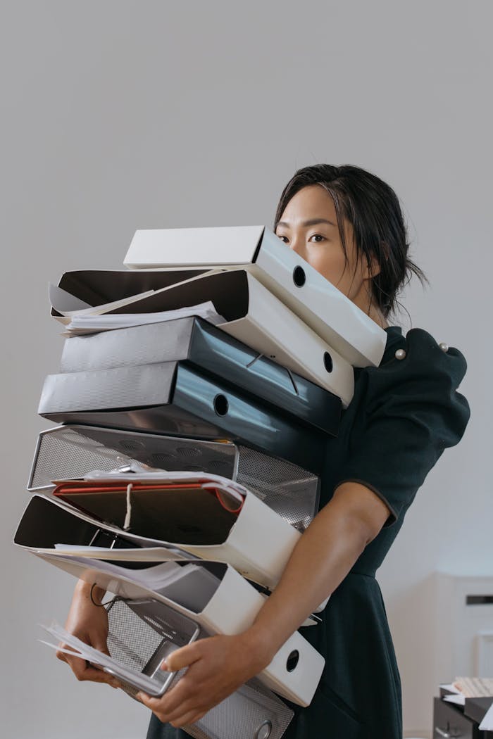 portfolio-01 Woman carrying a large stack of file folders indoors, expressing stress and workload.